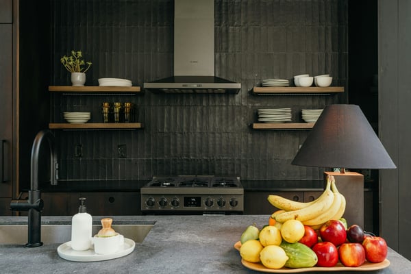 Modern kitchen with dark tile backsplash, wooden shelves holding dishware, gas range, black faucet, and gray countertop displaying fresh fruit and table lamp.