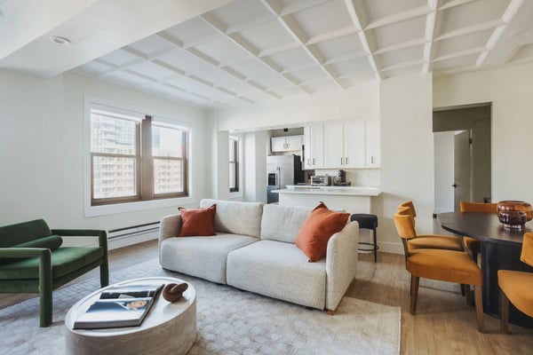 Open-plan living room with beige sofa, green and yellow chairs, exposed beam ceiling, and kitchen visible in background