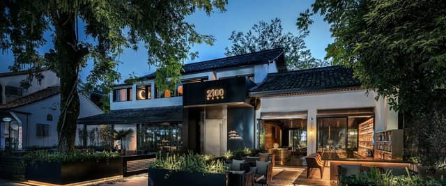 Modern home exterior at dusk with illuminated entrance, black awning, wooden deck, and mature trees surrounding white and dark facade.
