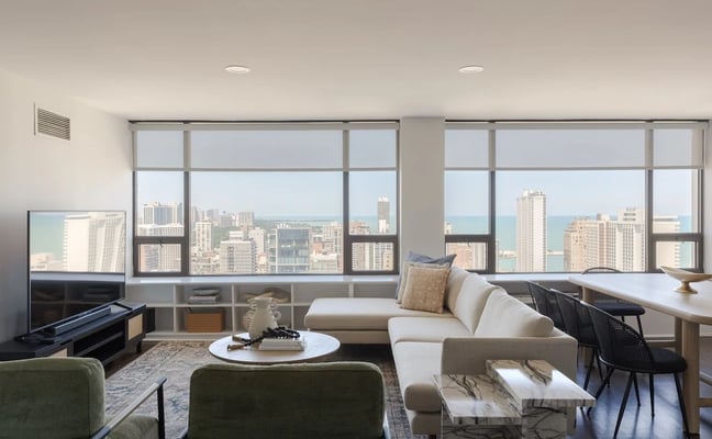 High-rise living room with floor-to-ceiling windows overlooking city skyline and lake, cream sofa, green chairs, and marble coffee table.