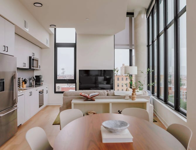 Modern open-plan apartment with white kitchen, dining table, living room with TV, and floor-to-ceiling windows overlooking city skyline.