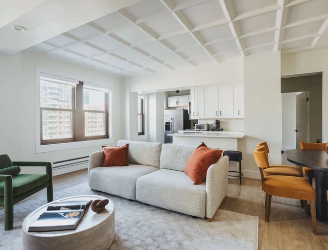 Modern open-plan living room with beamed ceiling, cream sofa, green and yellow chairs, and kitchen visible in background