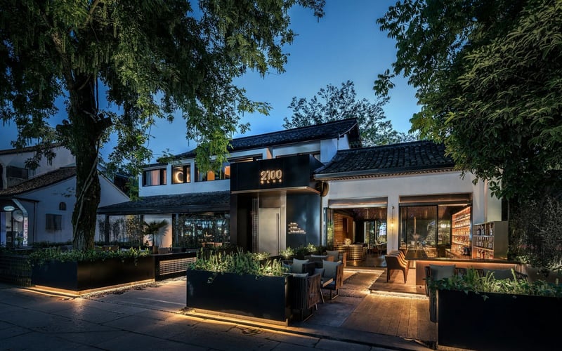 Modern house exterior at dusk with illuminated black storefront, white walls, dark roof, and mature trees surrounding the property